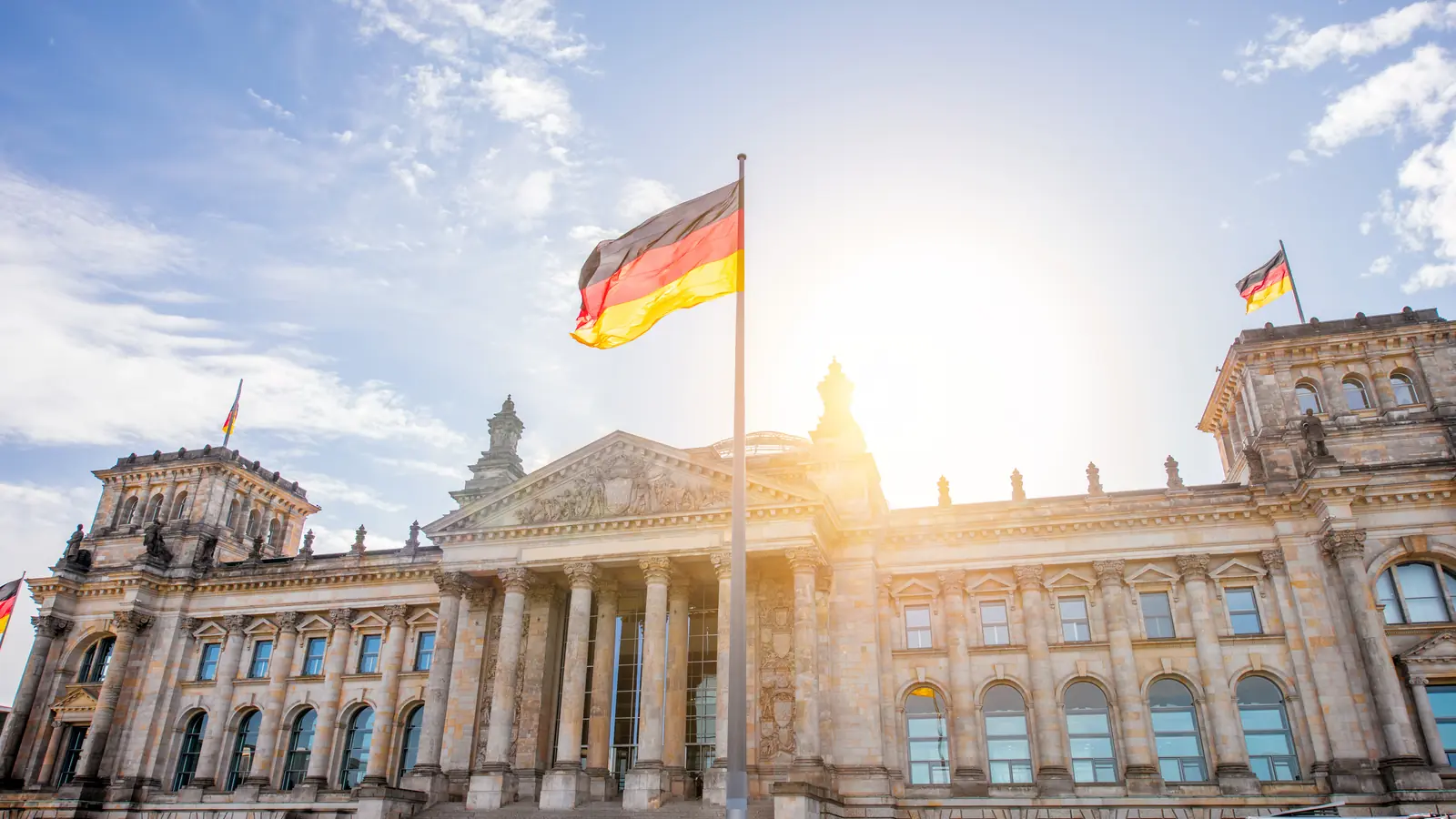 Toà nhà Quốc hội Reichstag ở Berlin với quốc kỳ Đức tung bay, mặt trời chiếu ngược phía sau.