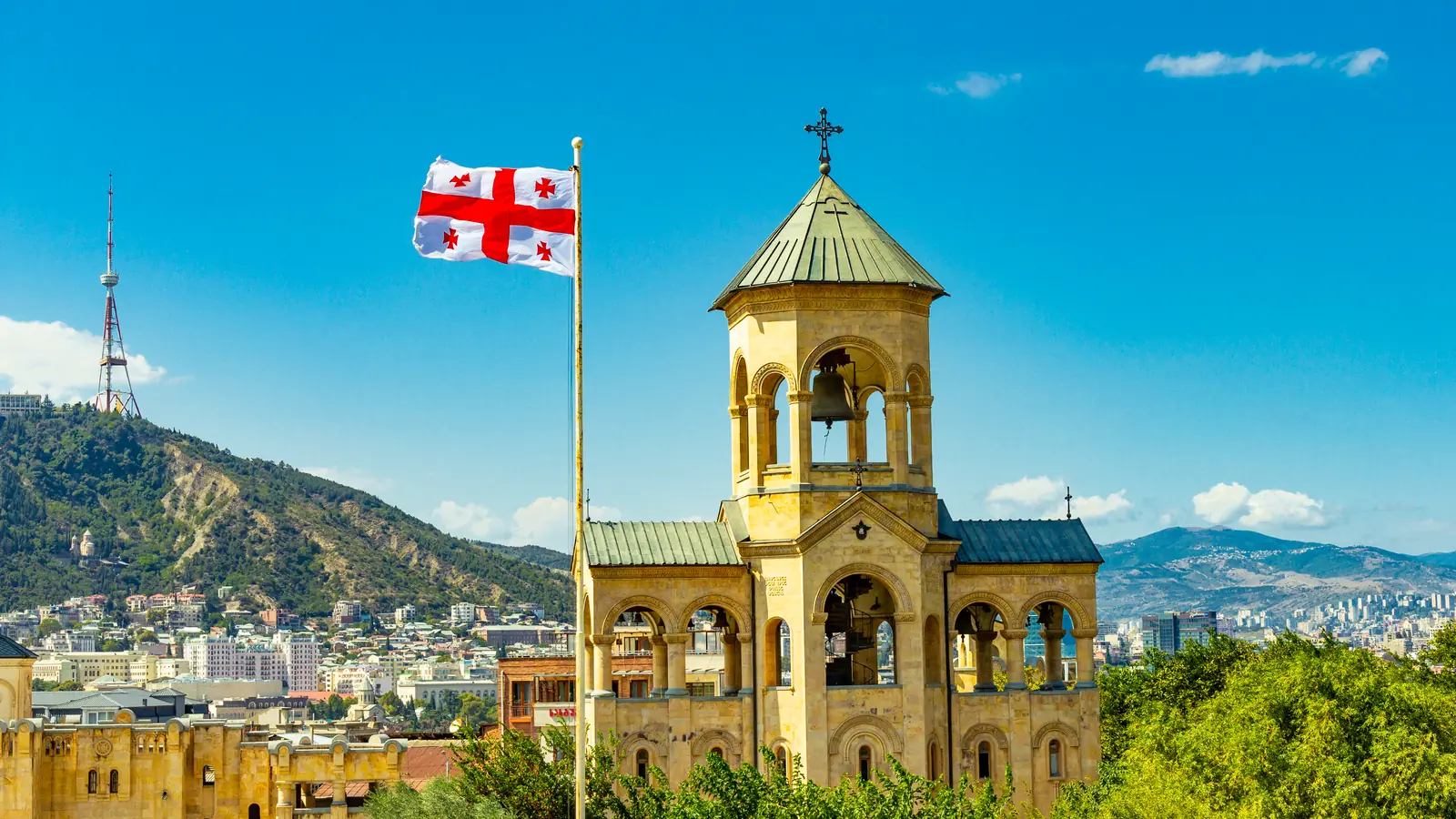 Tháp chuông nhà thờ bằng đá cạnh quốc kỳ Georgia trên cao nhìn xuống Tbilisi, tháp truyền hình trên ngọn đồi phía sau.