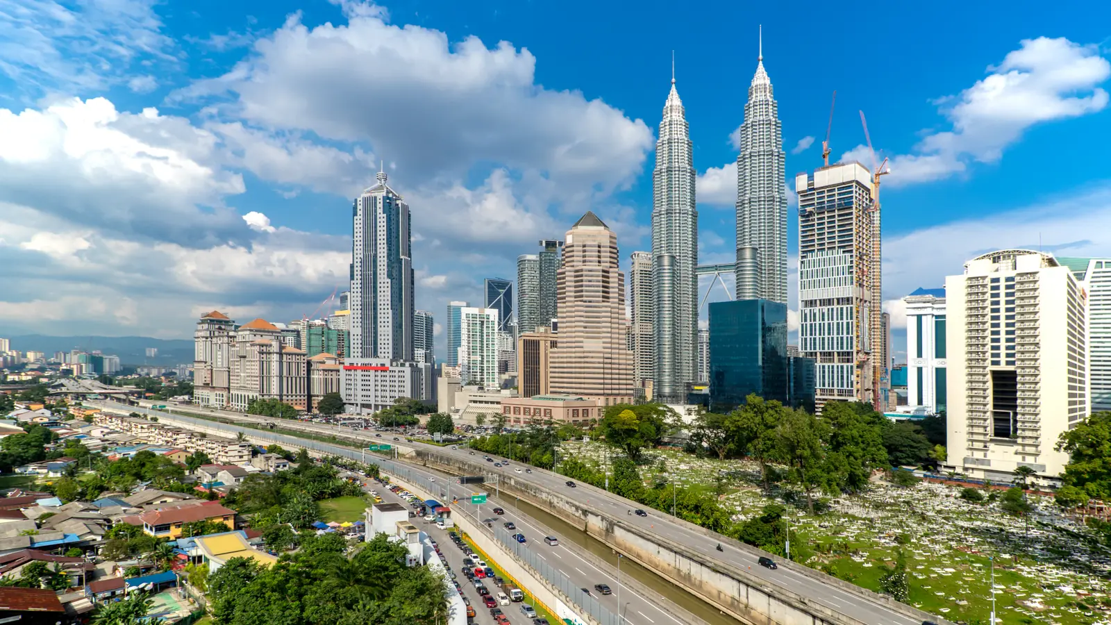 Tháp đôi Petronas và cụm cao ốc Kuala Lumpur dưới trời xanh mây trắng, đường cao tốc ở tiền cảnh.