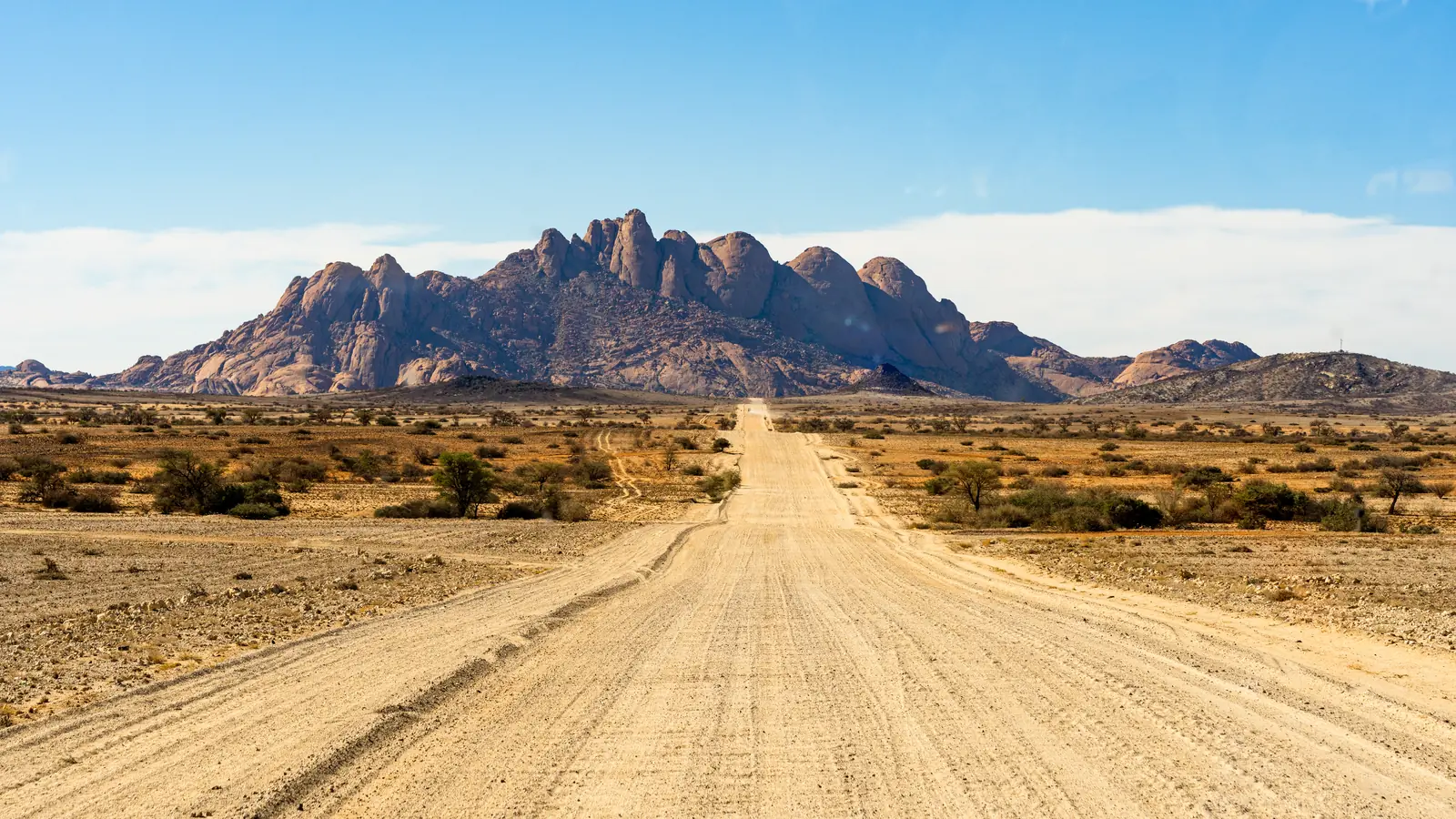 A straight gravel road across a dry plain leading towards a jagged granite massif.