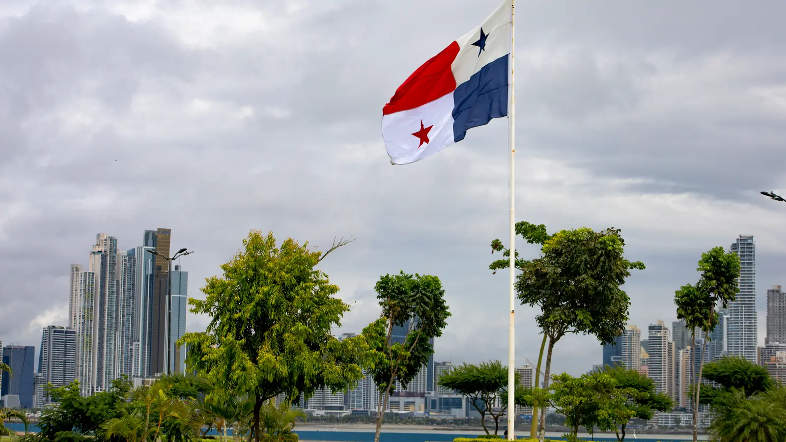 Quốc kỳ Panama bay trên cột cờ, cụm cao ốc Thành phố Panama sau hàng cây dưới trời nhiều mây.