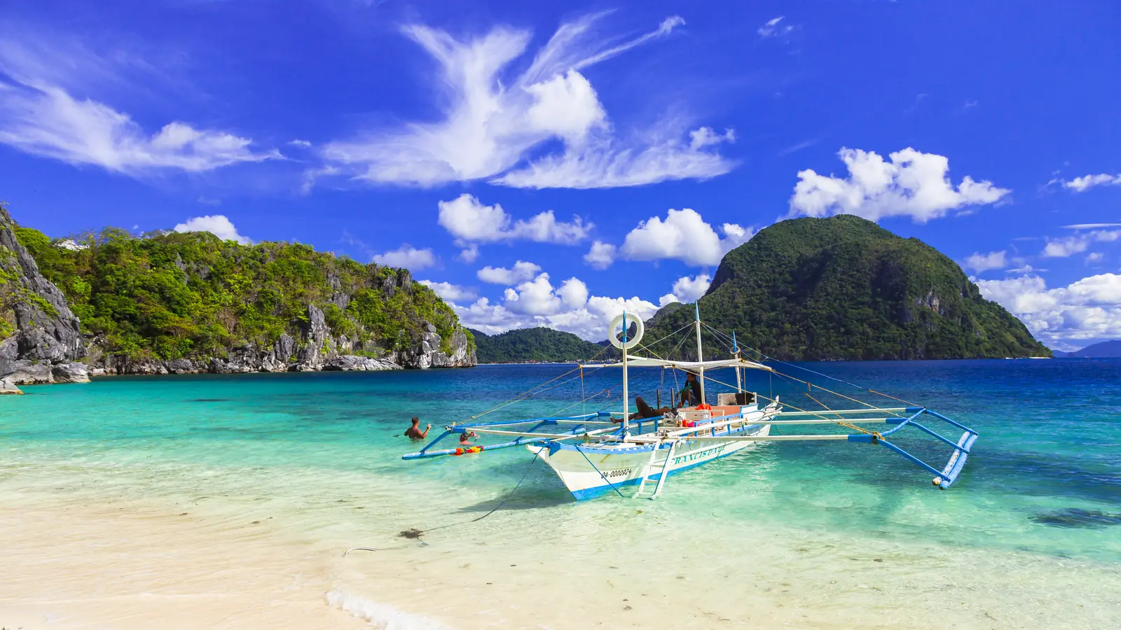 Thuyền gỗ có phao hai bên đậu trên bãi biển nước trong, các đảo đá vôi phủ cây xanh phía sau.