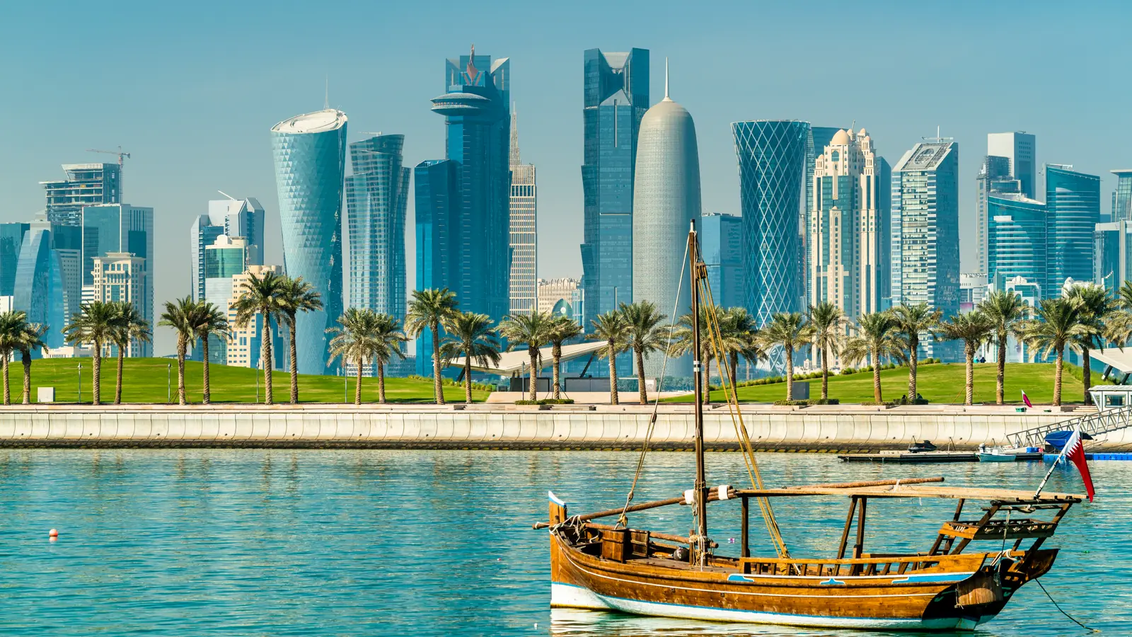 A traditional wooden dhow on Doha Bay, palm trees and the West Bay towers behind.