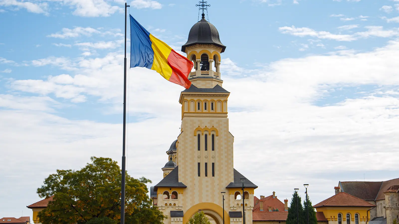 Quốc kỳ Romania bay cạnh tháp chuông của một nhà thờ chính toà dưới trời xanh mây trắng.