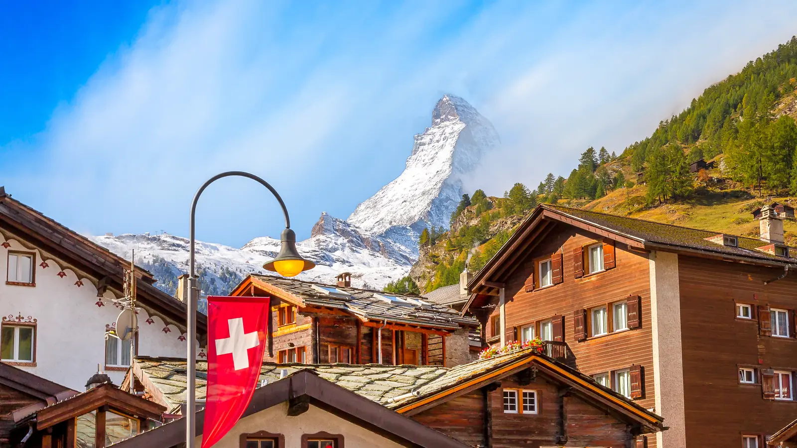 Đỉnh Matterhorn phủ tuyết phía sau những ngôi nhà gỗ ở Zermatt, quốc kỳ Thụy Sĩ treo trên cột đèn.