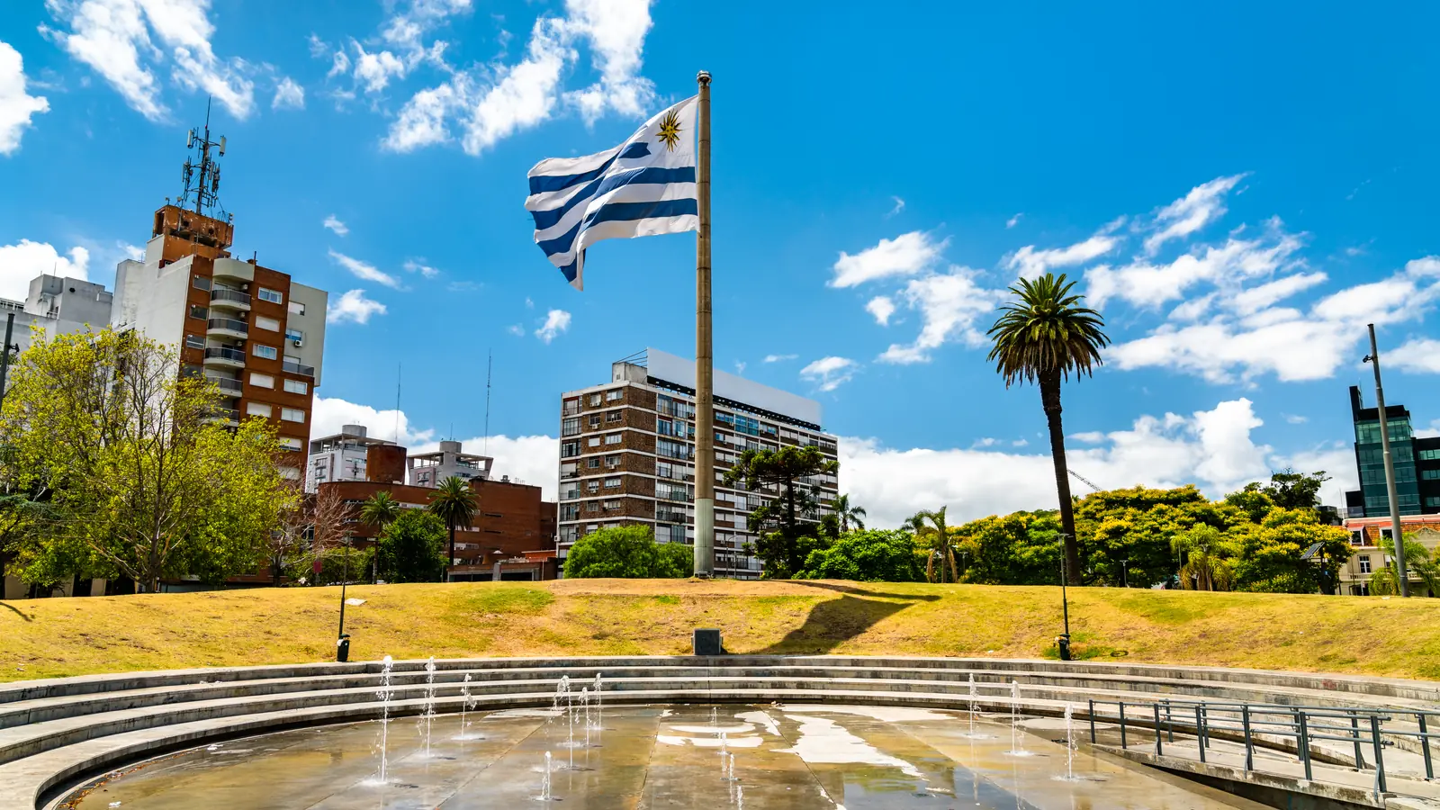 Quốc kỳ Uruguay bay trên cột cờ giữa quảng trường có đài phun nước, cây cọ và các toà nhà căn hộ xung quanh.