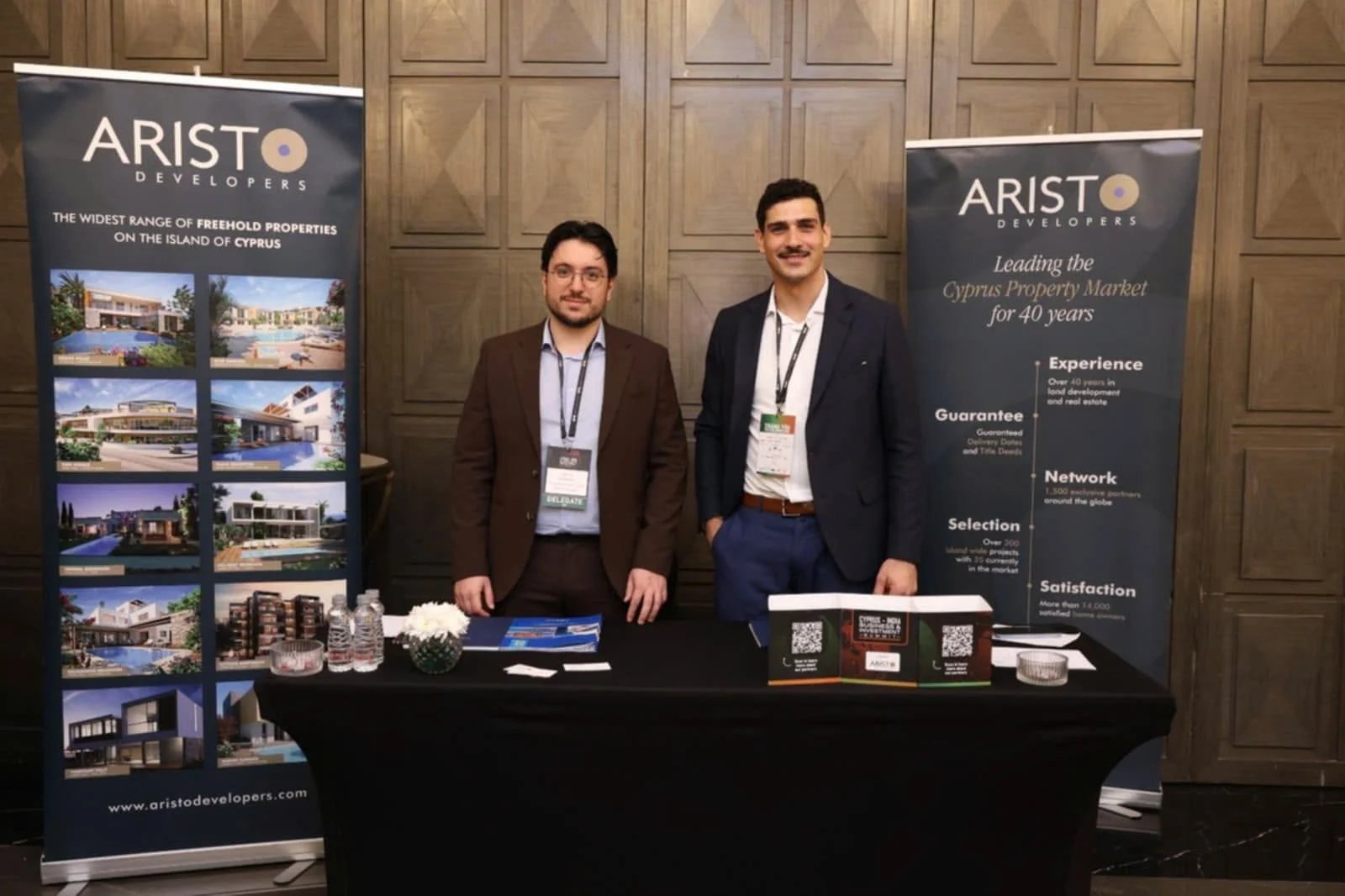 Hai người đàn ông mặc vest đứng sau bàn trưng bày phủ khăn đen, hai bên là hai băng rôn đứng Aristo Developers in ảnh dự án và các con số của công ty