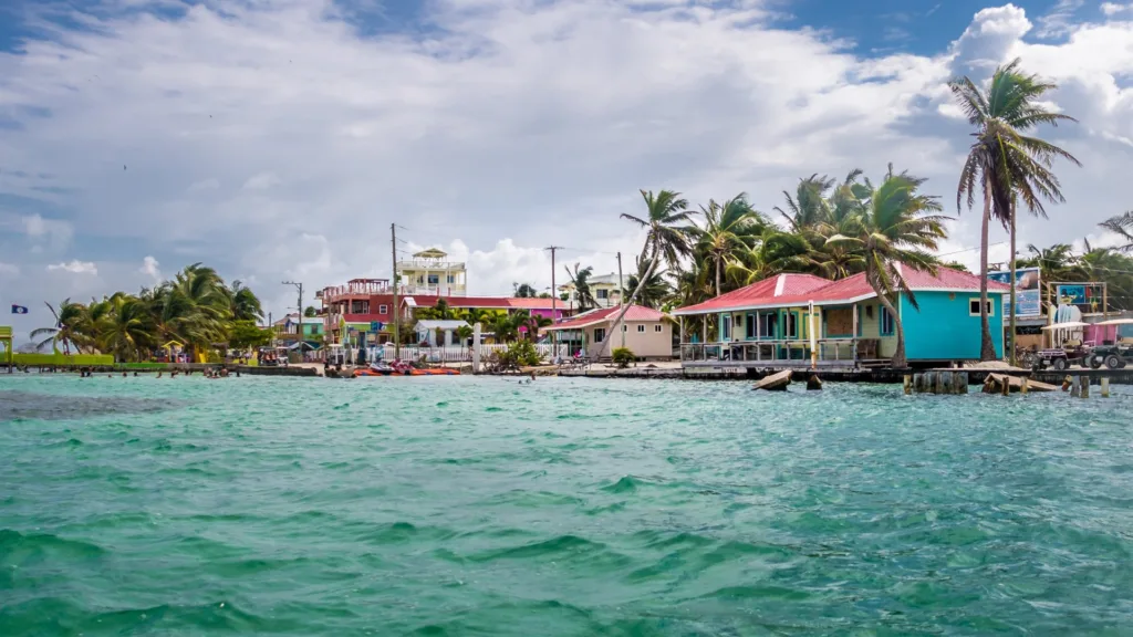 Painted timber houses on stilts along a shoreline with palm trees and small jetties, seen across shallow turquoise water under a cloudy sky.