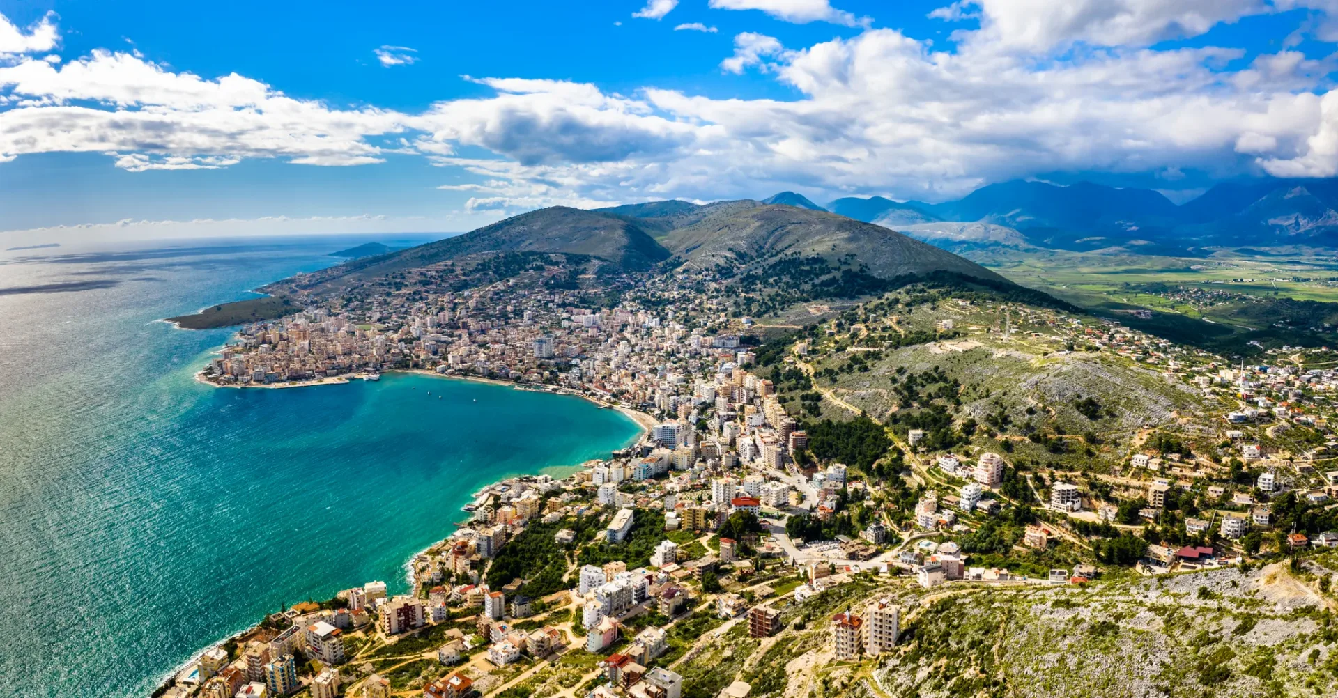 Saranda from above: the turquoise Ionian bay curving around a strip of white buildings, green hillsides and mountains behind, southern Albania.