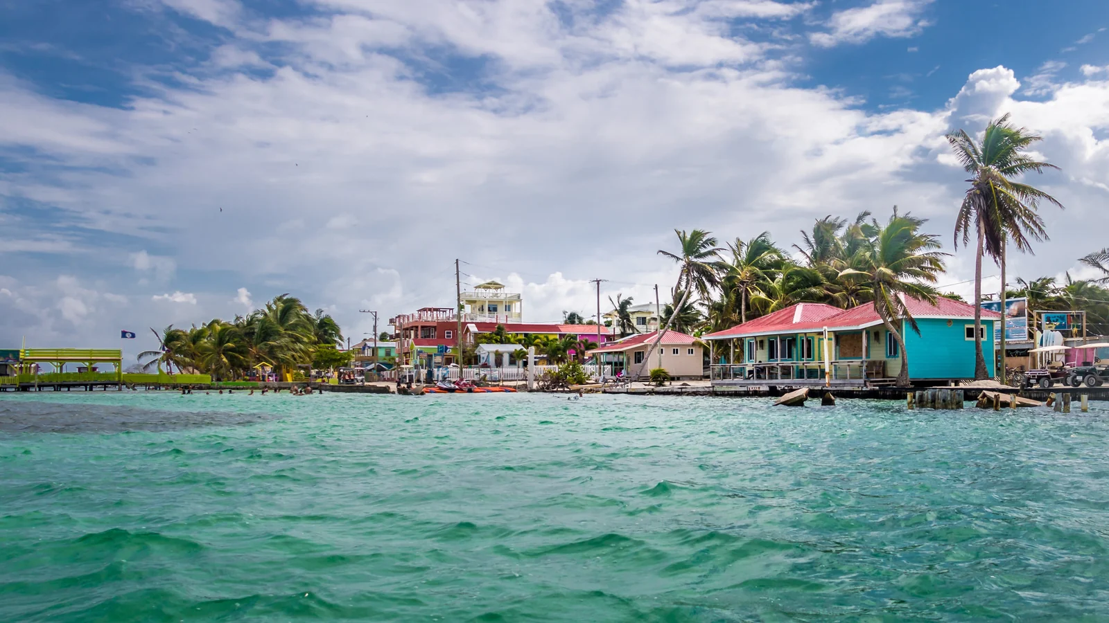 Xóm ven biển Belize với nhà gỗ sơn màu dựng trên cọc, hàng dừa và quốc kỳ Belize, nhìn từ mặt nước ngọc bích.