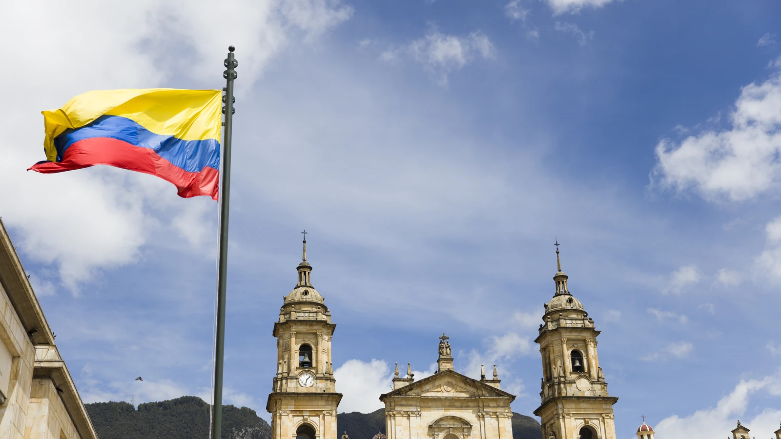 The Colombian flag flying in front of Bogotá's cathedral, its two bell towers and stone facade under a cloudy blue sky, hills behind.