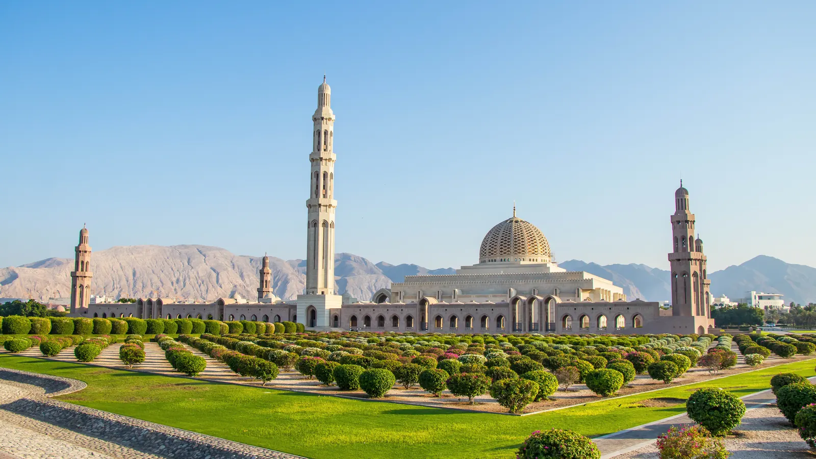 The Sultan Qaboos Grand Mosque seen from the gardens: a golden dome, one tall minaret at the centre and two shorter ones flanking it, against bare rocky mountains