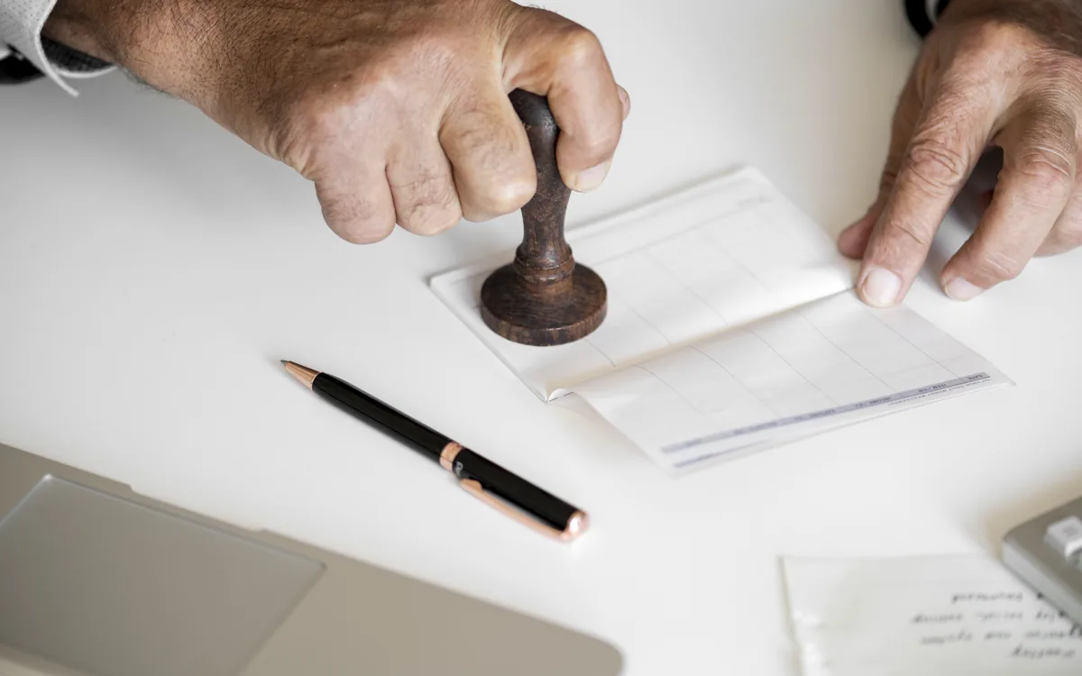 Two hands pressing a wooden seal stamp onto a stack of papers on a white table.