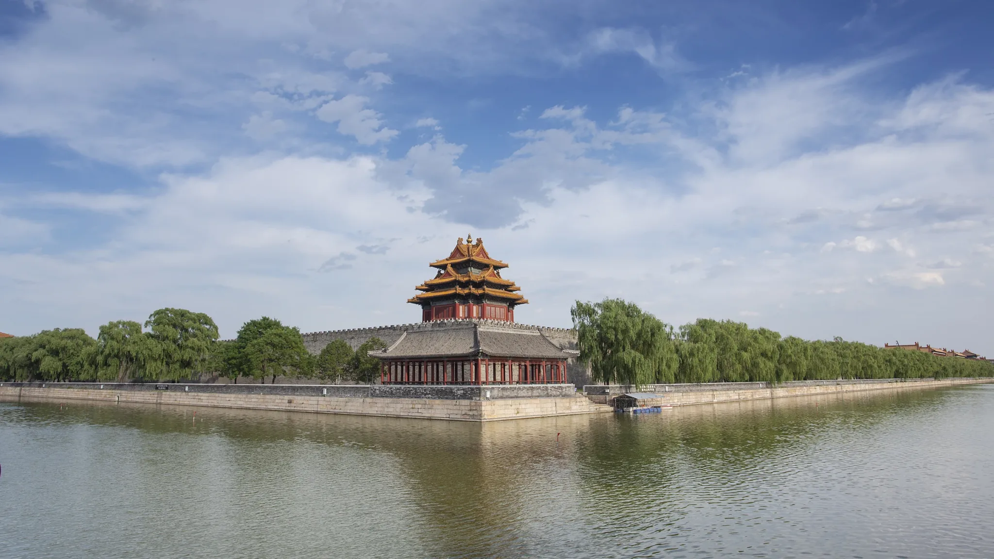 A curved-roof corner tower of the Forbidden City reflected in the moat, a row of willows along the wall behind.