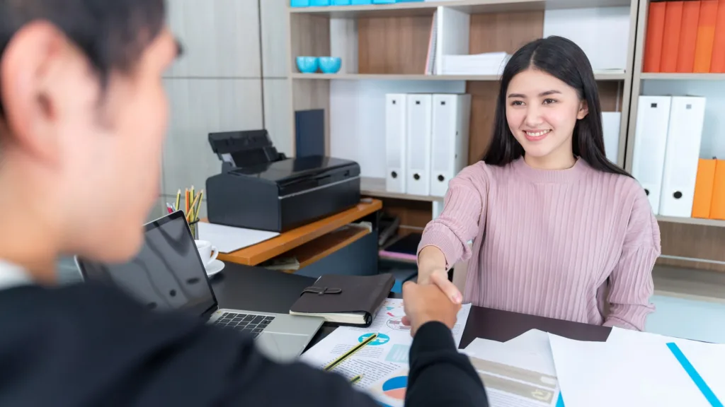 A woman behind a desk in an office shakes hands with a client seated opposite her, with printed charts and a laptop on the desk between them.