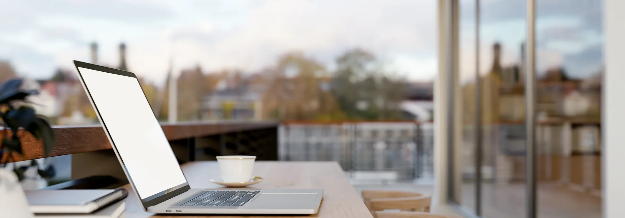 An open laptop on a wooden table beside a glass window, with rooftops and a town bell tower outside.