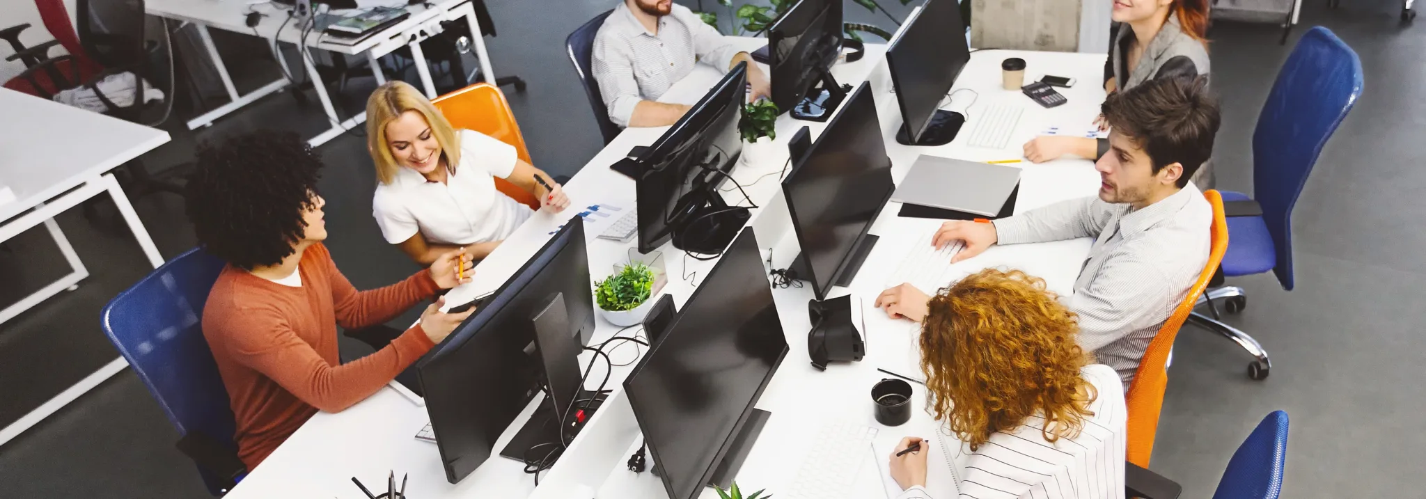 A group of people seated around two joined rows of desks in a shared office, computer monitors down the middle, seen from above.