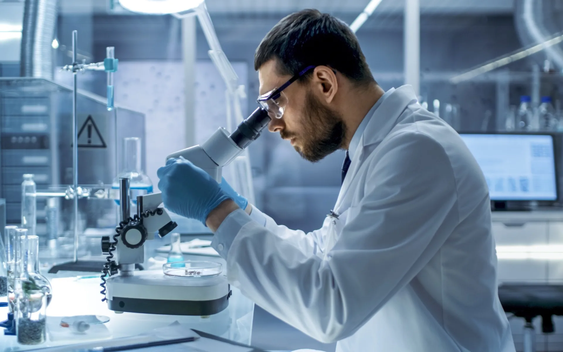 A researcher in a white lab coat, safety glasses and blue gloves looks into a microscope beside a Petri dish in a laboratory