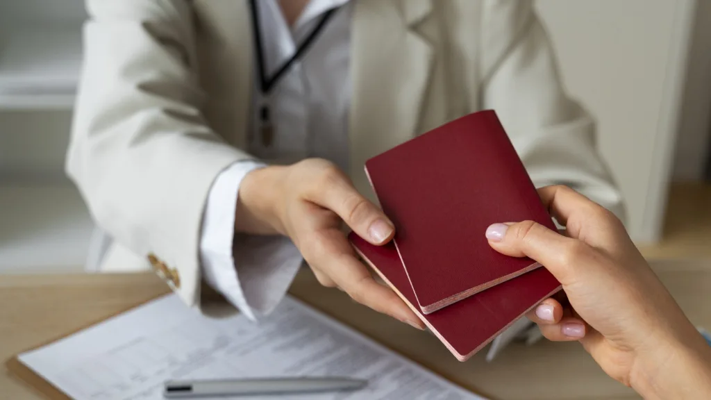 An official passes two passports across a desk to an applicant, beside a clipboard holding a completed form and a pen.