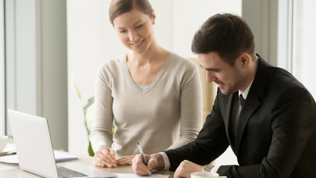 Một phụ nữ và một người đàn ông ngồi tại bàn làm việc, ký vào một tờ giấy, trên bàn có máy tính xách tay và tách cà phê.