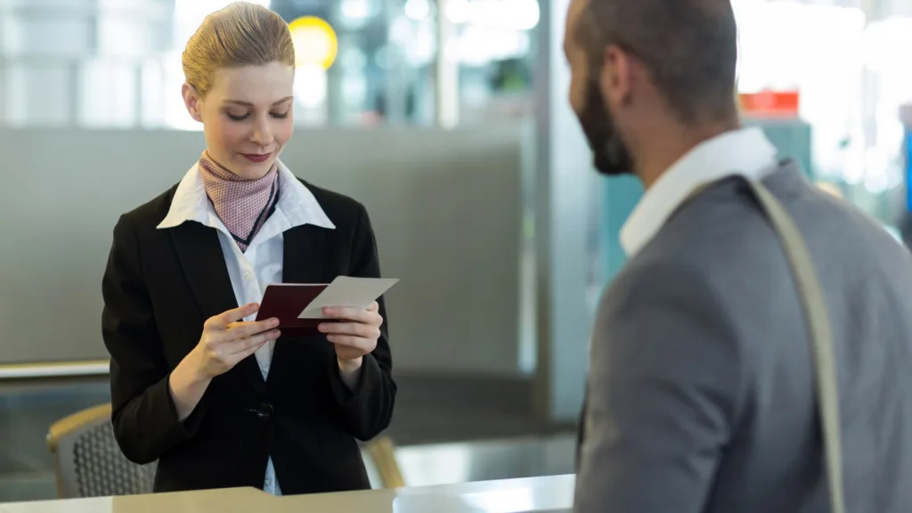 A counter attendant examines a passport while a traveller in a grey jacket waits.