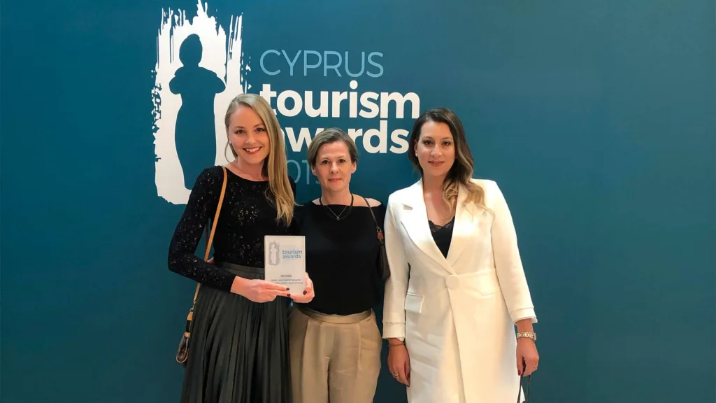 Ba người đứng trước phông nền xanh đậm in chữ Cyprus Tourism Awards, người đứng giữa cầm một kỷ niệm chương bằng thuỷ tinh.
