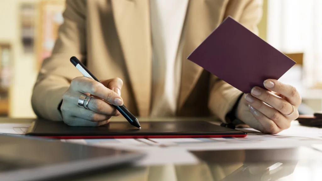 A person in a jacket signs on a tablet with one hand while holding a plain burgundy passport in the other.