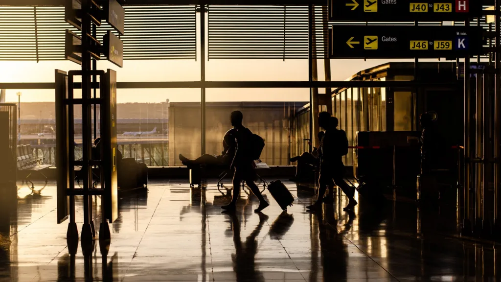 Two travellers with wheeled suitcases cross a glazed airport concourse in silhouette at sunset, beneath overhead signs pointing to departure gates.