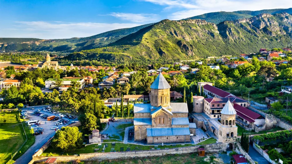 Aerial view of a stone church with a grey conical dome and monastery buildings among trees, below green mountains in Mtskheta, Georgia