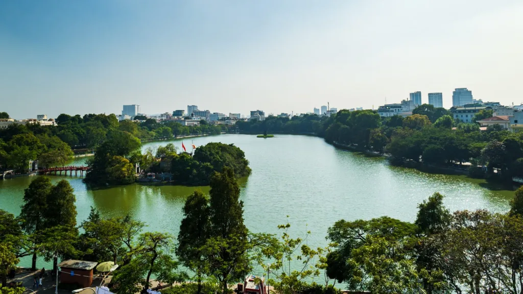 High view over Hoan Kiem Lake in Hanoi, with the red Huc Bridge, a wooded island, trees along the shore and city buildings beyond