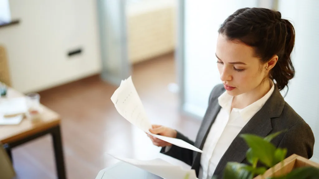 A woman in a dark blazer stands turning through a sheaf of printed pages, reading one of them. The text on the pages is not legible. An office with a wooden floor is out of focus behind her.