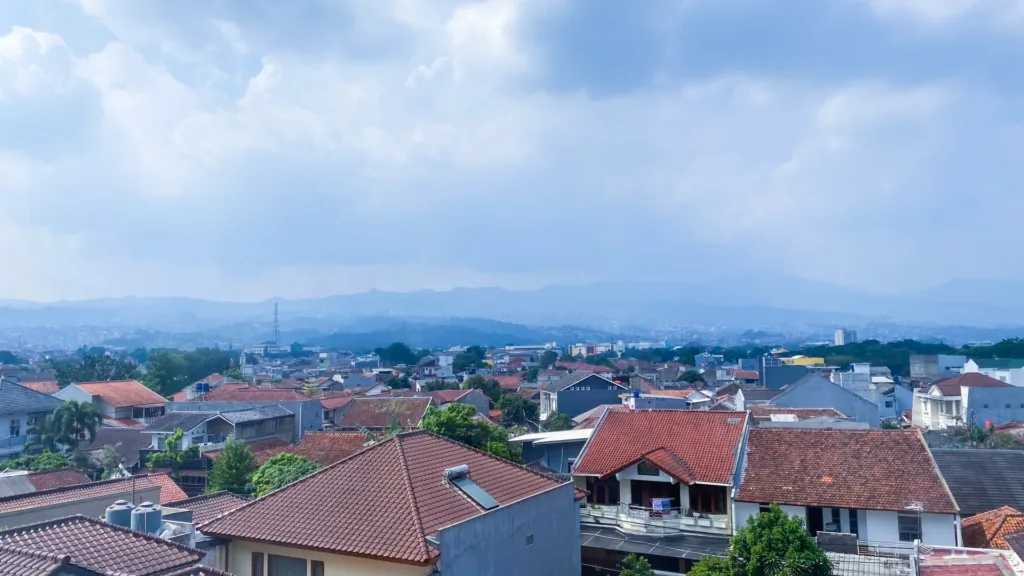 Tiled rooftops of a residential district in Bandung, Indonesia, seen from above, with hills and heavy cloud behind.