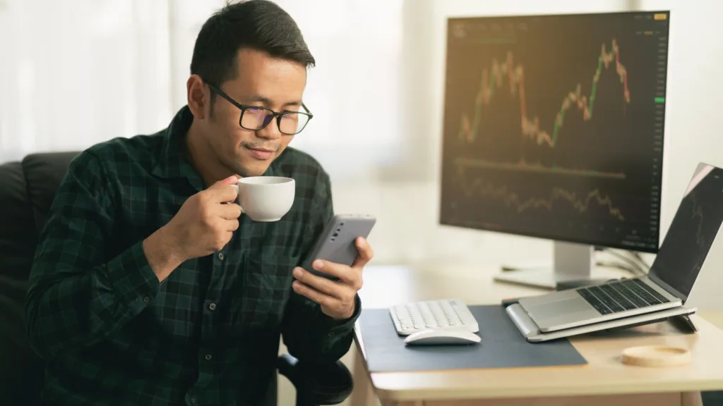 Một người đàn ông đeo kính cầm tách cà phê, xem điện thoại tại bàn làm việc, phía sau là màn hình hiển thị biểu đồ giá
