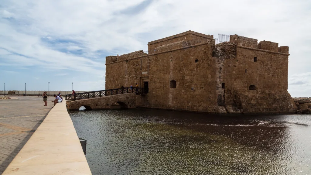 Lâu đài cảng Paphos xây bằng đá bên mặt nước, có cầu dẫn vào và vài du khách đứng trên bờ kè