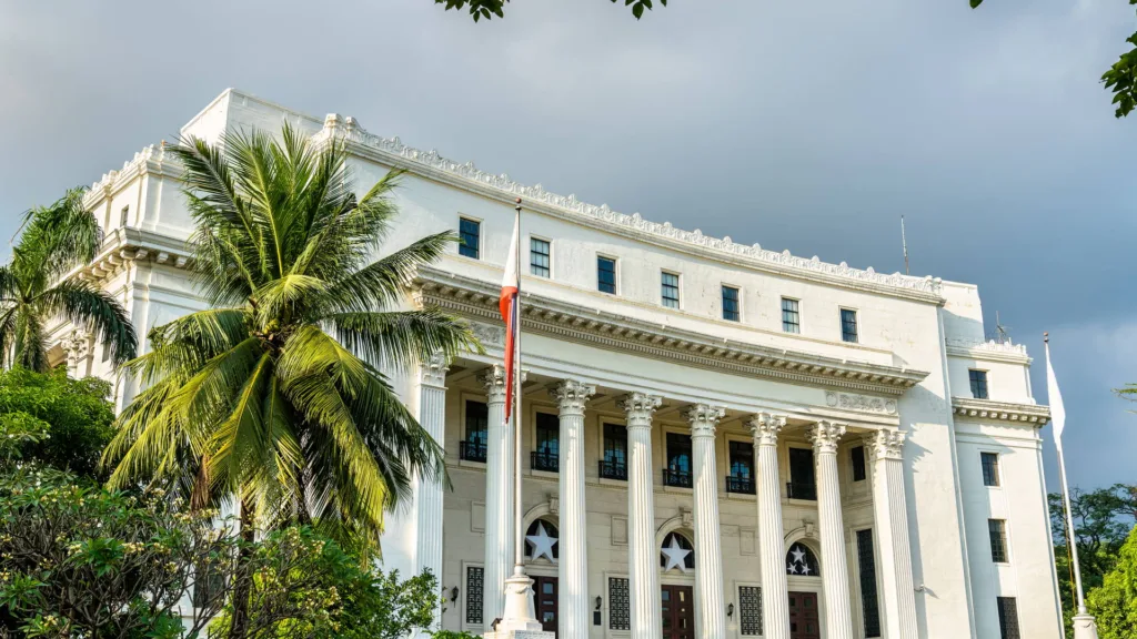 A white neoclassical public building in Manila photographed from below, with Corinthian columns across the portico, the Philippine flag hanging from a mast at the centre, and a coconut palm in the foreground.