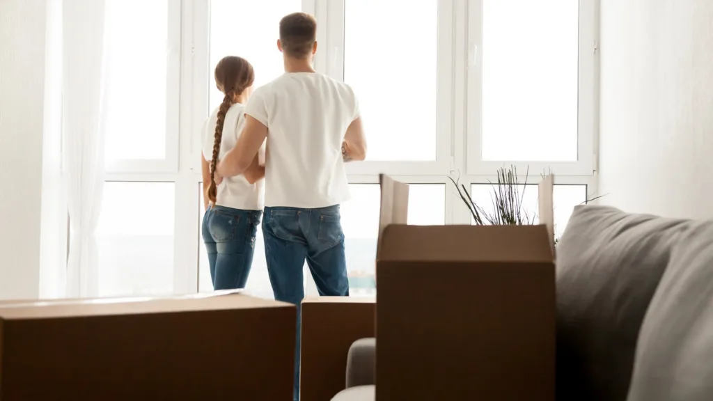 Một cặp đôi đứng nhìn ra cửa sổ lớn trong căn hộ mới, quanh họ là những thùng các-tông chưa mở