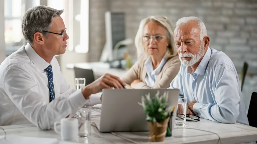 An adviser in a shirt and tie points to a laptop screen at a table, facing an older man and woman who are watching the screen. Papers, a water glass and a small plant sit on the table.