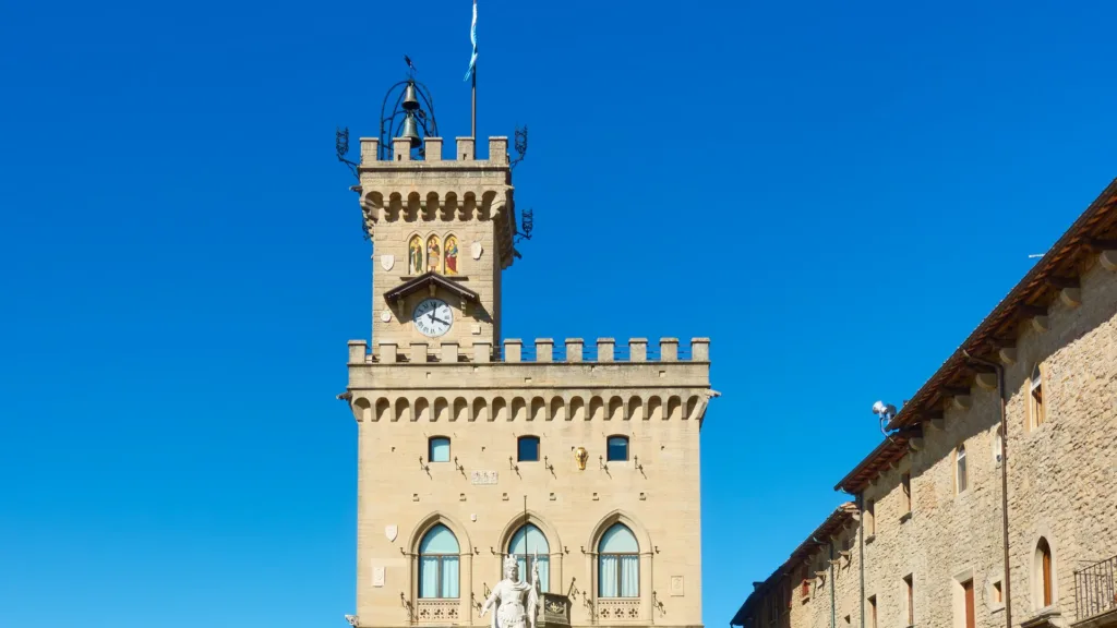 Mặt tiền đá có lỗ châu mai và tháp đồng hồ của Palazzo Pubblico ở San Marino dưới trời xanh trong, phía trước là một bức tượng trắng.