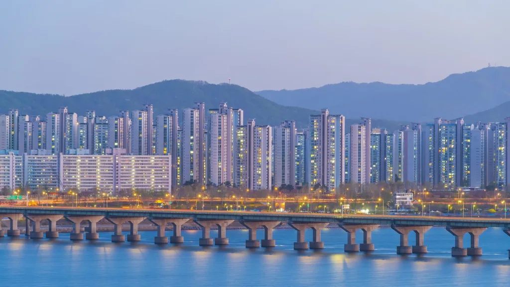 Rows of high-rise apartment towers at dusk behind a long bridge over the Han River in Seoul, with mountains in the background
