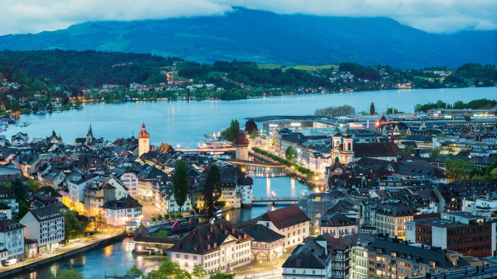 Aerial view at dusk of a lakeside old town with lit streets and church towers, a river flowing into the lake and wooded hills behind, in Lucerne, Switzerland