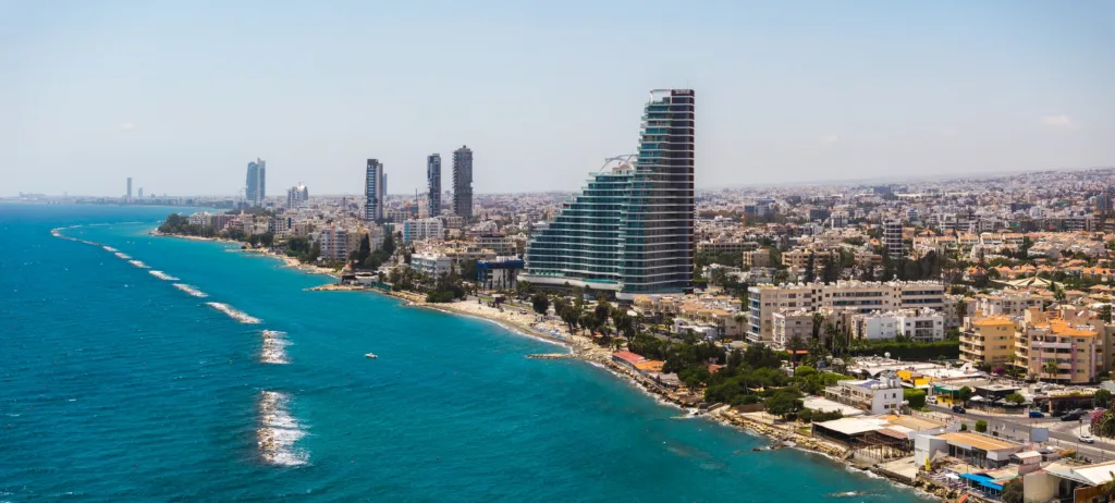 Bờ biển Limassol nhìn từ trên cao: Dãy nhà cao tầng và một toà tháp kính uốn cong chạy dọc bãi biển, bên làn nước Địa Trung Hải xanh ngọc