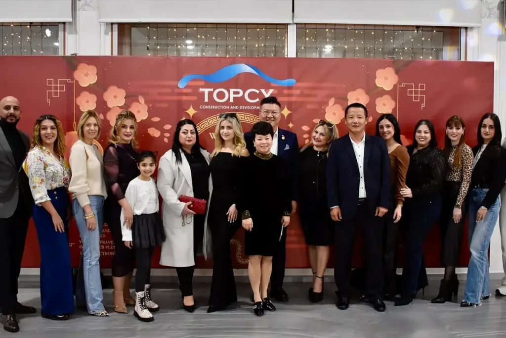 Khoảng hai mươi người đứng thành hàng trước phông đỏ in logo TOPCY trang trí hoa mai