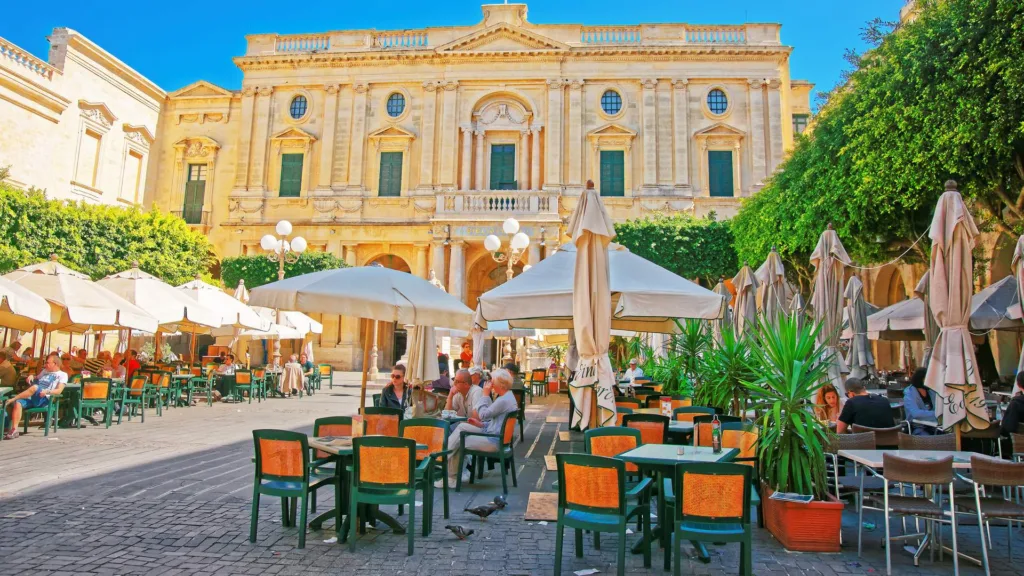 Cafe tables under parasols in Republic Square, Valletta, in front of the National Library of Malta.