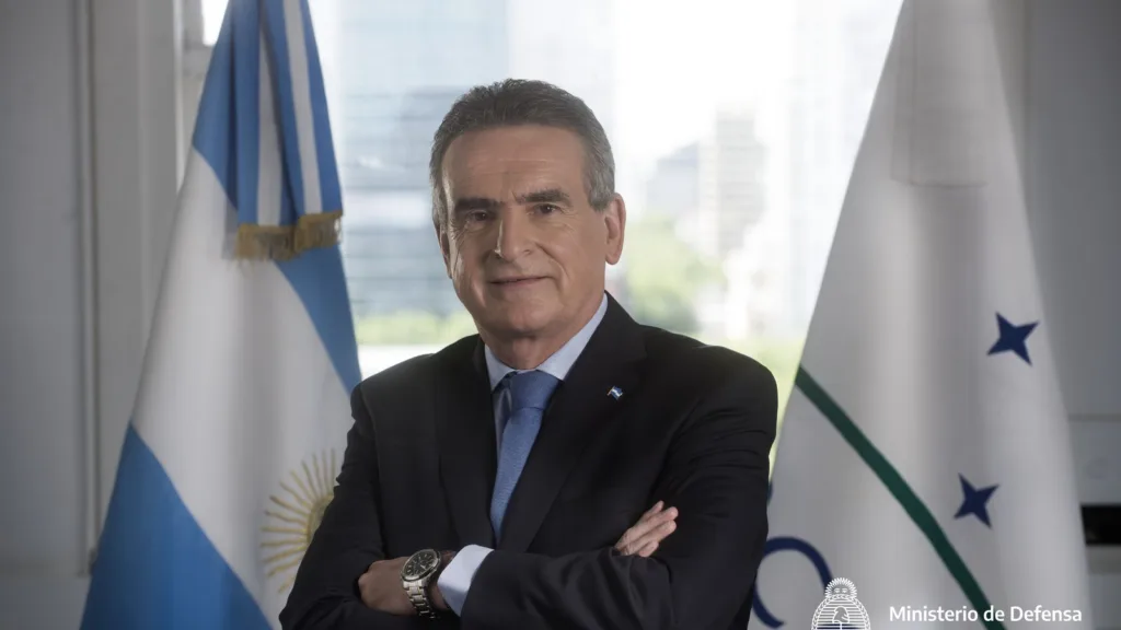 Agustín Rossi in a dark suit and blue tie, arms folded, standing between the Argentine flag and a Mercosur flag
