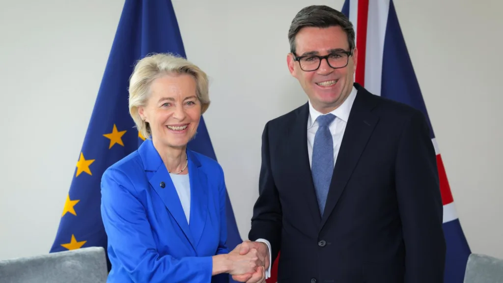 Bà Ursula von der Leyen mặc áo khoác xanh bắt tay ông Andy Burnham trước cờ EU và cờ Anh