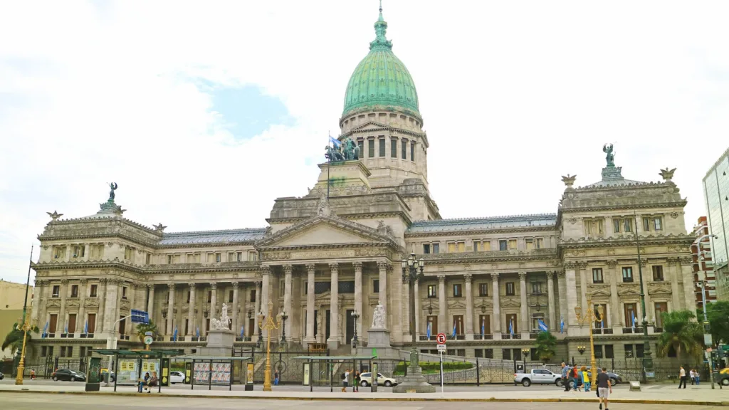 The green-domed Palace of the Argentine National Congress in Buenos Aires seen from across the avenue, with pedestrians and bus shelters in the foreground and blue-and-white flags along the facade
