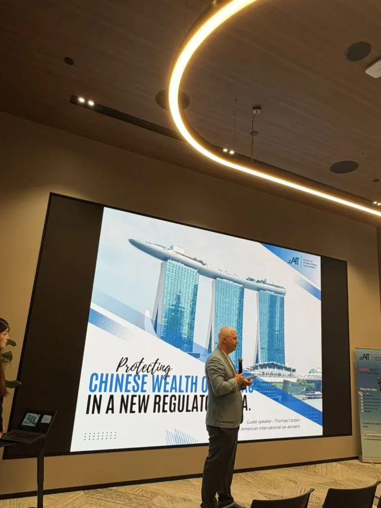 Một diễn giả đứng trước màn hình lớn chiếu trang trình bày "Protecting Chinese Wealth Overseas in a New Regulatory Era" có hình Marina Bay Sands