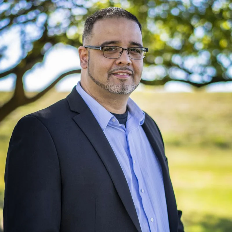 Portrait of Milton Barboza wearing glasses, a dark jacket and a blue shirt outdoors