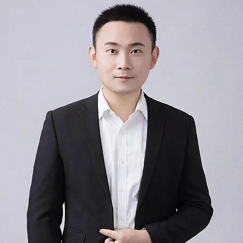 Portrait of Qi Zhou in a black jacket and white shirt against a light grey background