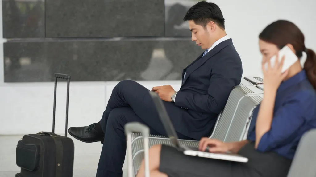 A man in a navy suit sits on a perforated metal bench in a terminal waiting area beside a black wheeled suitcase, while a woman in the foreground types on a laptop and talks on the phone